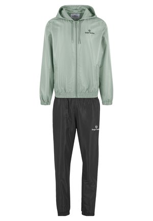Homme debout portant une veste zippée à capuche vert clair, un pantalon noir et des baskets blanches sur un fond clair uni.