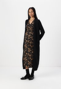 Lungo cardigan nero indossato sopra un vestito nero con motivo floreale dorato. Il modello indossa calzini neri e scarpe con punta affusolata con dettagli borchiati.