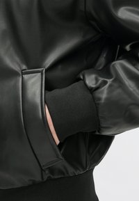 Mustmust must jacket, millemust viimistlusega, millel on ribatud must varrukas ja tasku detail varrukal.