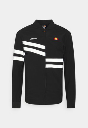 Veste noire zippée avec des rayures blanches diagonales et horizontales, logo ellesse sur le côté gauche de la poitrine et nom de la marque sur l'épaule droite.