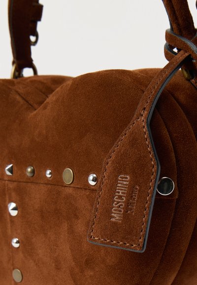 Sac à main en suède marron de forme arrondie, avec des clous en métal et une étiquette cousue embossée "MOSCHINO Milano" sur la sangle.