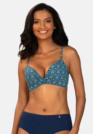 Kobieta w niebieskim wzorzystym bikini z cienkimi ramiączkami i jednokolorowych, ciemnoniebieskich majtkach bikini, uśmiechająca się na jednolitym tle.