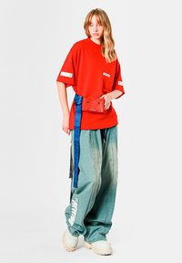 Camiseta roja extragrande con logo, vaqueros anchos azul claro y zapatos crema con plataforma. Incluye un cinturón azul y una riñonera roja.