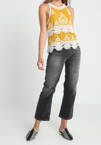 Keltainen pitsireunainen hihaton toppi, jossa on pyöreä pääntie, yhdistetty mustiin suoraan lahkeeseen tuleviin farkkuihin, joissa on polven repeämä, sekä ruudulliset liukusandaalit.