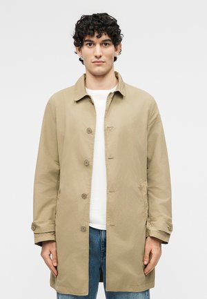 Νεαρός άνδρας με σγουρά μαλλιά που φοράει μπεζ παλτό με κουμπιά πάνω από λευκό πουκάμισο και μπλε τζιν, στέκεται μπροστά σε απλό φόντο.