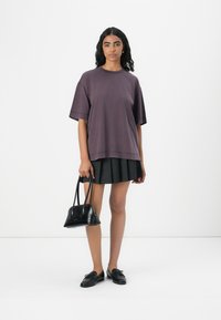 T-shirt viola oversize a maniche corte, abbinato a una gonna nera a pieghe. Tiene in mano una piccola borsa nera. Mocassini neri completano il look.