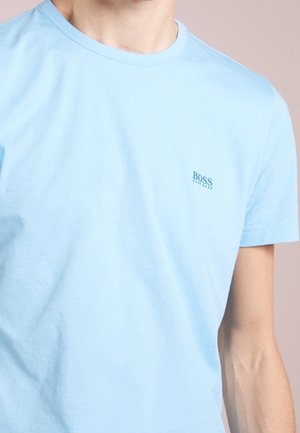 Mężczyzna ubrany w jasnoniebieską koszulkę z krótkim rękawem i okrągłym dekoltem marki BOSS, obciętą przy szyi i górnej części ramienia.