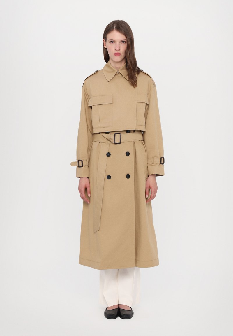 Cappotto trench beige con vita cintrata e cintura, bottoni doppi, e grandi tasche con patta. Presenta un colletto classico e cinturini ai polsini.