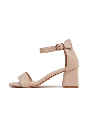 Beige Wildleder-Sandalen mit Absatz, geschlossenem Fersenbereich, breitem Riemen über den Zehen und verstellbarem Knöchelriemen mit goldener Schnalle.