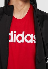 Svart zip-up jacka delvis öppen, som avslöjar en röd T-shirt med stort vitt "adidas"-logotyp. Jackan har en strukturerad krage och detaljer.