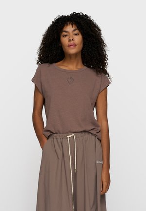 Kobieta o kręconych włosach, ubrana w lawendowy top bez rękawów i luźną beżową spódnicę ze sznurkiem, stojąca na jasnym, jednolitym tle.