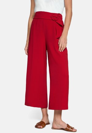 Femme portant un pantalon rouge taille haute à jambes larges et courtes, avec ceinture en tissu et boucle, assorti à des sandales plates marron et un haut blanc.