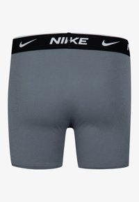 Pelēki kokvilnas bokseršorti ar melnu elastīgu jostu, kurai ir balts "NIKE" teksts un logo. Gluda tekstūra, pieguļošs siluets un bez redzamām rakstām.