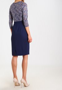Femme portant une robe bleu marine ajustée avec des manches trois-quarts en dentelle et des chaussures à talons hauts couleur nude, debout de dos sur fond blanc.