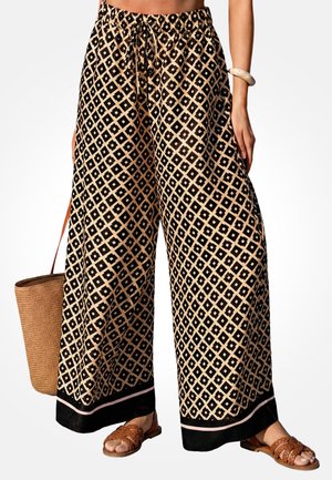 Luźne spodnie o szerokich nogawkach z beżowo-czarnym geometrycznym wzorem diamentowym, noszone z brązowymi plecionymi sandałami i plecioną torbą na ramię.