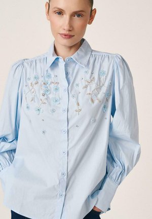 Mujer con blusa azul claro con mangas abullonadas y delicado bordado floral en tonos azules y beige en el pecho.