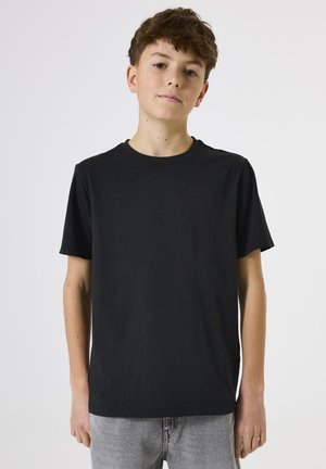 Tienerjongen met kort bruin haar, gekleed in een effen zwart T-shirt met korte mouwen en lichtgrijze jeans, staand tegen een witte achtergrond.