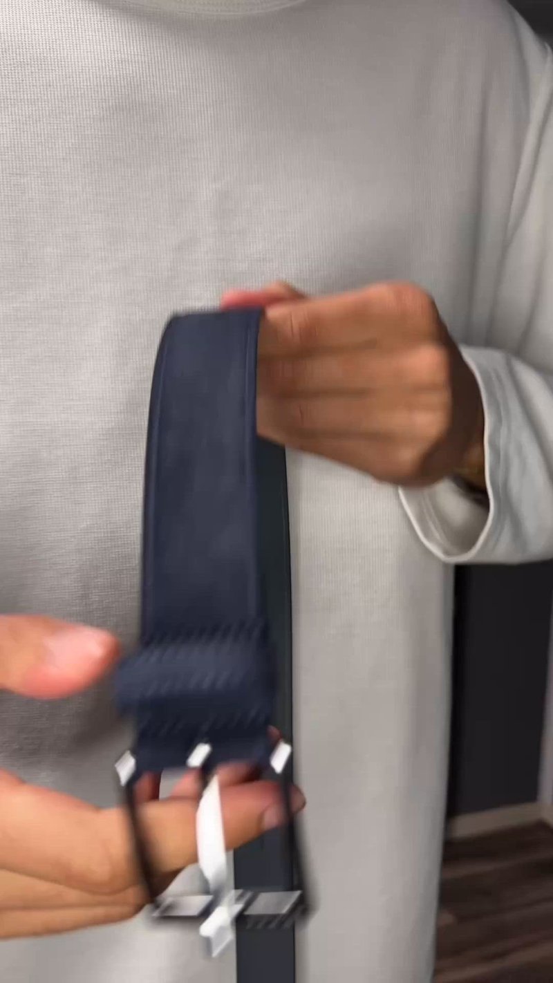 Homme portant un pull blanc tenant une ceinture bleu marine avec une boucle argentée devant un fond sombre.