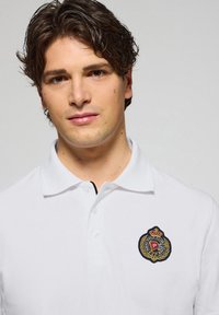 Witte polo shirt gemaakt van textuurstof, met een marineblauwe ronde geborduurde badge met gouden accenten op de linkerkant van de borst.