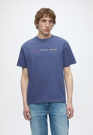 Jeune homme portant un t-shirt bleu « Tommy Jeans » avec un texte rouge, blanc et jaune, associé à un jean bleu clair, debout devant un fond blanc.