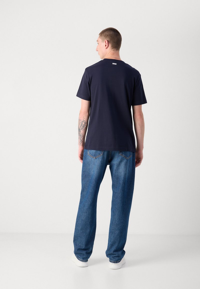 Granatowa bawełniana koszulka z krótkim rękawem i okrągłym dekoltem, noszona z jasnoniebieskimi dżinsami. Metka widoczna w okolicy tyłu dekoltu.