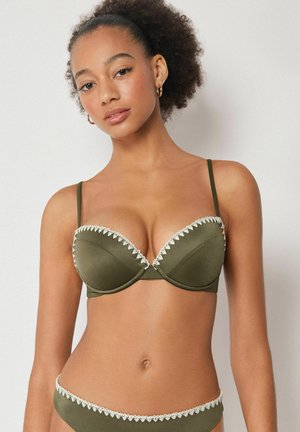 Frau mit lockigem Haar, die einen olivgrünen BH und passende Unterwäsche mit weißer Spitzenverzierung trägt, frontal vor einfarbigem Hintergrund blickend.
