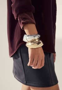 Drei klobige metallische Armreifen in Gold, Silber und mit reflektierendem Finish, getragen an einem Handgelenk, kombiniert mit einem burgunderroten Hemd und einem schwarzen Lederrock.