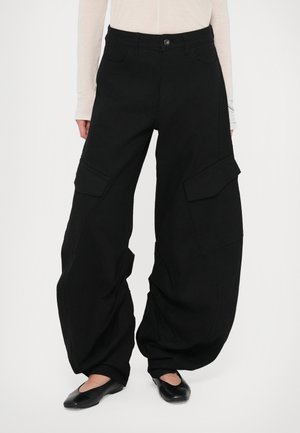 Calças cargo pretas com um corte solto, apresentando bolsos laterais, fecho de botão e um tecido texturizado. Usadas com sapatos planos pretos.