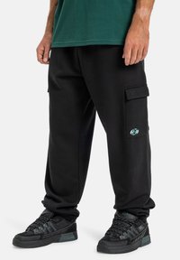 Schwarze Cargo-Hosen mit seitlichen Taschen, die ein kleines Logo aufweisen. Tragen Sie sie mit strukturierten schwarzen Sneakers und stehen Sie vor einem neutralen Hintergrund.