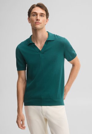 Jonge man met een teal korte mouwen polo en witte broek, staand met één hand achter zijn rug tegen een effen achtergrond.