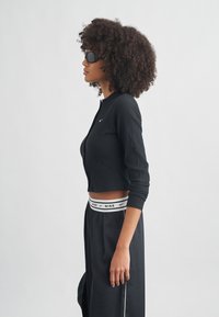 Μαύρο ριμπ crop top με μακριά μανίκια, με μικρό λογότυπο της Nike. Συνδυάζεται με μαύρο παντελόνι που διαθέτει λευκή ζώνη μέσης με το λογότυπο της Nike.
