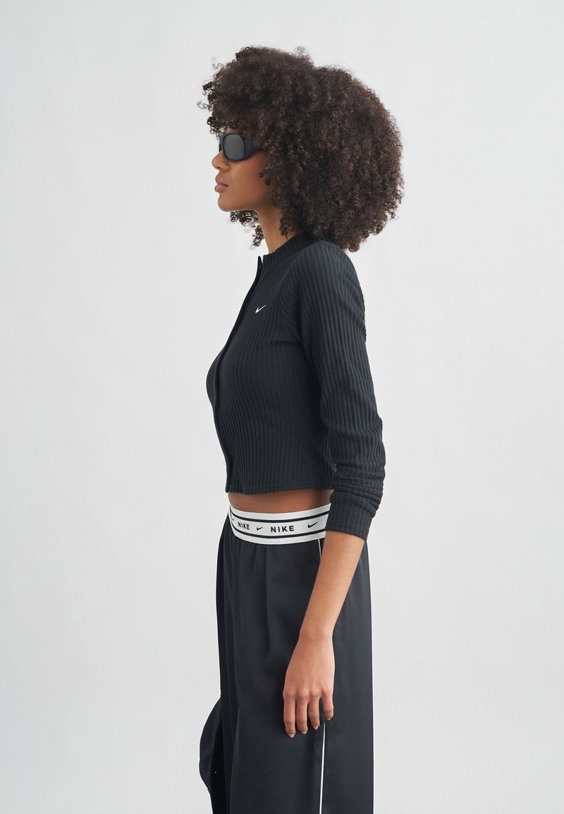 Μαύρο ριμπ crop top με μακριά μανίκια, με μικρό λογότυπο της Nike. Συνδυάζεται με μαύρο παντελόνι που διαθέτει λευκή ζώνη μέσης με το λογότυπο της Nike.
