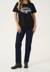 Černé tričko s krátkým rukávem s grafickým motivem rtů ze stříbrných flitrů, doplněné tmavě modrými džínami a černými kotníkovými botami.