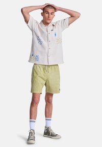 Joven con pelo corto está de pie con las manos en la cabeza, vistiendo una camisa de manga corta estampada, pantalones cortos verdes, calcetines blancos y zapatillas grises de caña alta.