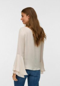 Blusa color beige chiaro con maniche svasate e una consistenza liscia. La parte posteriore presenta un collo arrotondato e una vestibilità comoda, abbinata a jeans blu.