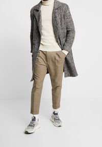 Grå prickig kappa, krämfärgad polo, beige avsmalnande byxor och grå sneakers med vita sulor. Avslappnad, lager-på-lager stil.