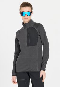 Grau melierter Pullover mit Reißverschluss, schwarzer Brusttasche, langen Ärmeln und strukturiertem Stoff. Das Modell trägt reflektierende blaue Sonnenbrillen.