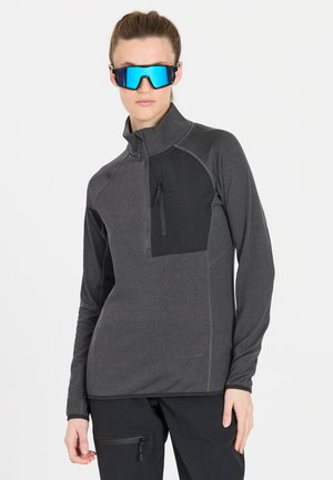 Grå lynlåset pullover med en sort brystlomme, lange ærmer og tekstureret stof. Modellen bærer reflekterende blå solbriller.