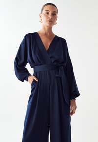 Marineblauwe jumpsuit in wikkelstijl met lange, losse mouwen, een ceintuur in de taille en zakken. Gladde stof met een lichte glans.