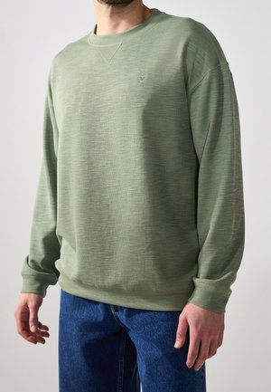 Mand iført lys grøn langærmet rundhalset sweater og blå jeans, stående foran en ensfarvet lys baggrund.