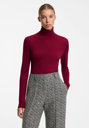 Mujer con un suéter ajustado de cuello alto burdeos y pantalones de talle alto con patrón espiga gris, con la mano izquierda en el bolsillo.