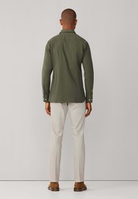 Langarmig olijfgroen shirt van gladde stof met een knoopsluiting bij de kraag, gecombineerd met lichtgekleurde broek en bruine schoenen.