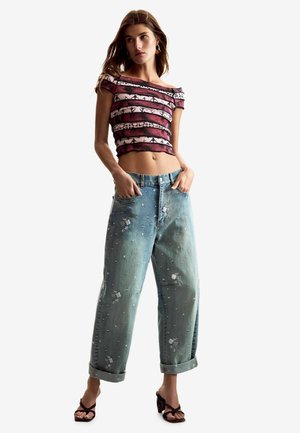 Naine, kes kannab roosat mustrilist õlgadeta lühikest topi, lahtisi helesiniseid tikanditega pükse, mille mansetid on üles keeratud, ja musti avatud varbaga kontsakingi.