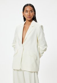 Blazer de lino blanco con solapa de muesca, cierre de un solo botón y dos bolsillos frontales. Presenta un corte relajado y un diseño ligeramente extragrande.