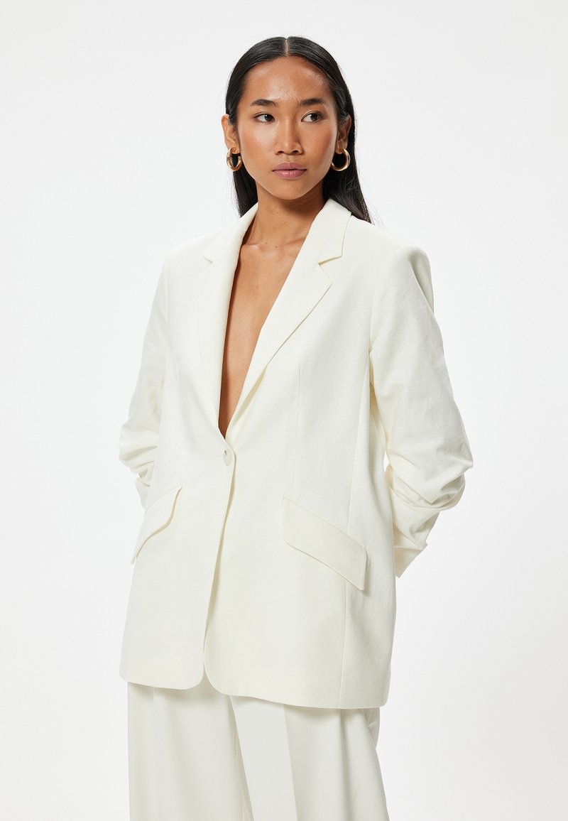 Blazer de lino blanco con solapa de muesca, cierre de un solo botón y dos bolsillos frontales. Presenta un corte relajado y un diseño ligeramente extragrande.
