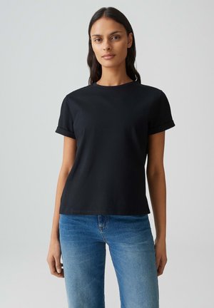 Vrouw met lang donker haar, gekleed in een eenvoudige zwarte T-shirt met korte mouwen en een blauwe spijkerbroek, staand tegen een lichtgrijze achtergrond.