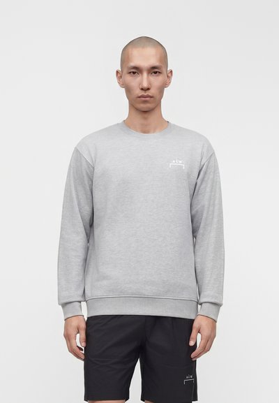 Homme à la tête rasée portant un sweat-shirt gris clair à manches longues et un short noir, debout devant un fond blanc uni.