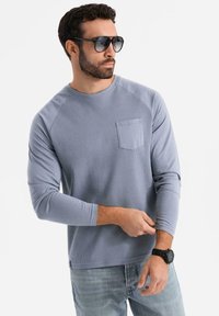 Vaalea sininen pitkähihainen paita, jossa on teksturoitu kangas, raglan-hihat ja rintatasku. Yhdistetty farkkujensa ja mustan kellon kanssa.