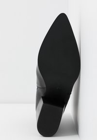 Chaussure noire à talon haut et bout pointu, posée sur son talon contre un mur blanc d'angle et un sol.