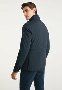 Granatowa kurtka wykonana z lekkiego materiału, z wysokim kołnierzem, bocznymi kieszeniami i logo na rękawie. Gładka tekstura, nowoczesny design.